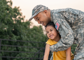 A smiling service member in camouflage hugs a smiling young child in a yellow shirt outdoors.