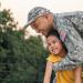A smiling service member in camouflage hugs a smiling young child in a yellow shirt outdoors.