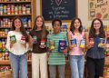 Five McGuffey Middle School students hold canned goods and boxed food while standing in front of stocked food pantry shelves.