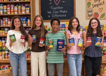 Five McGuffey Middle School students hold canned goods and boxed food while standing in front of stocked food pantry shelves.