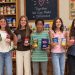 Five McGuffey Middle School students hold canned goods and boxed food while standing in front of stocked food pantry shelves.