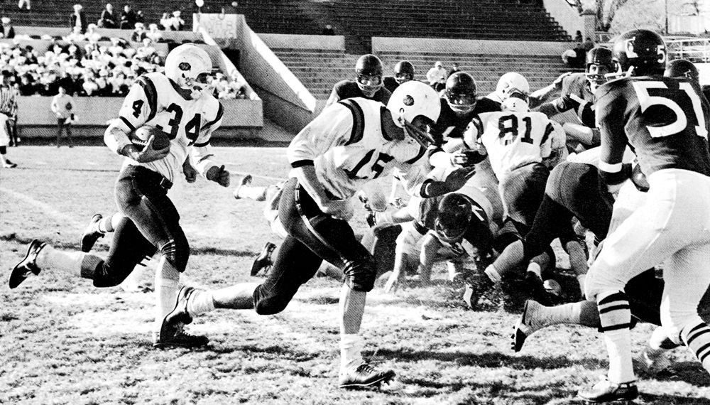 A vintage black-and-white photograph shows Waynesburg College football players in white uniforms running the ball against opposing defenders during a 1960s football game.