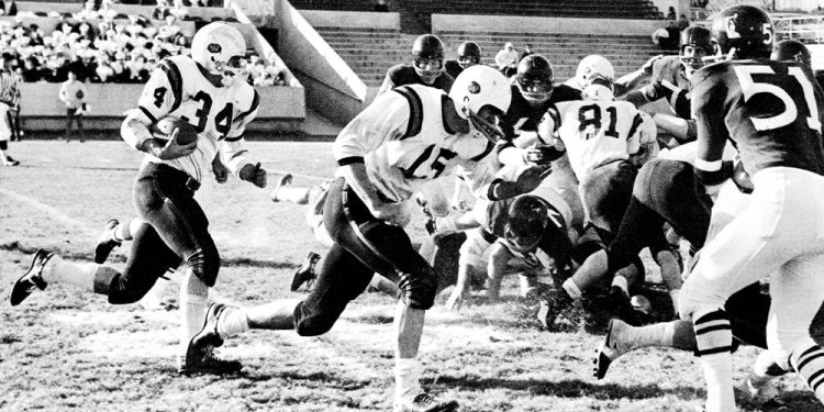 A vintage black-and-white photograph shows Waynesburg College football players in white uniforms running the ball against opposing defenders during a 1960s football game.