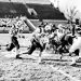 A vintage black-and-white photograph shows Waynesburg College football players in white uniforms running the ball against opposing defenders during a 1960s football game.