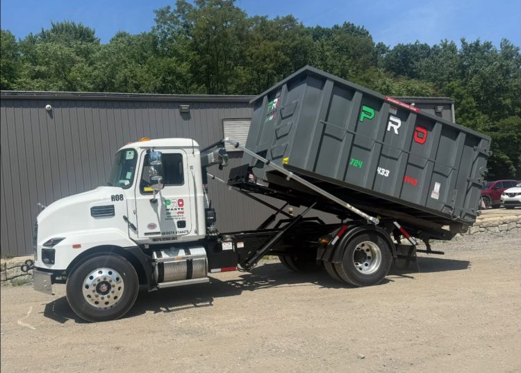 A white Pro Waste LLC truck with a raised gray roll-off dumpster is parked outside a commercial building in southwestern Pennsylvania.