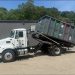 A white Pro Waste LLC truck with a raised gray roll-off dumpster is parked outside a commercial building in southwestern Pennsylvania.