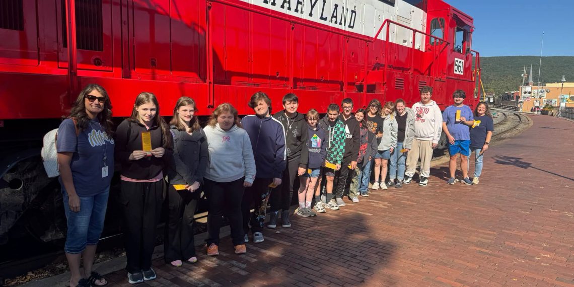 Carmichaels students and a teacher stand beside a red and white Western Maryland Railway locomotive during a fall field trip aboard the Frostburg Flyer.
