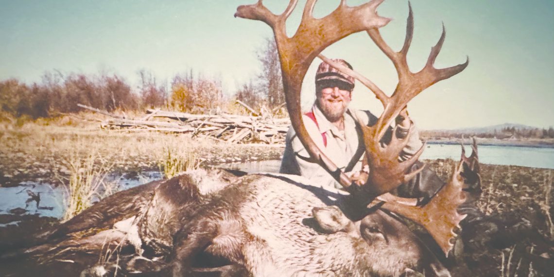 Hunter Larry Goehring poses behind a harvested bull caribou with massive antlers in a remote wilderness landscape near a body of water.