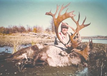 Hunter Larry Goehring poses behind a harvested bull caribou with massive antlers in a remote wilderness landscape near a body of water.