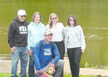 Aaron Houser kneels beside Coalbie, a therapy dog wearing a pink vest, as four supporters stand behind them along the Monongahela River in Greensboro, Pennsylvania.