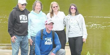 Aaron Houser kneels beside Coalbie, a therapy dog wearing a pink vest, as four supporters stand behind them along the Monongahela River in Greensboro, Pennsylvania.