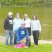 Aaron Houser kneels beside Coalbie, a therapy dog wearing a pink vest, as four supporters stand behind them along the Monongahela River in Greensboro, Pennsylvania.