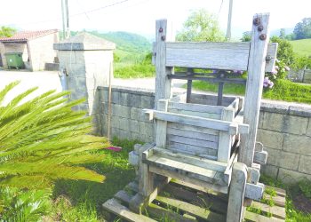 An antique wooden fruit press sits outdoors beside a stone wall in a scenic rural setting, illustrating traditional cider-making methods.
