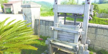 An antique wooden fruit press sits outdoors beside a stone wall in a scenic rural setting, illustrating traditional cider-making methods.
