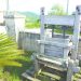 An antique wooden fruit press sits outdoors beside a stone wall in a scenic rural setting, illustrating traditional cider-making methods.