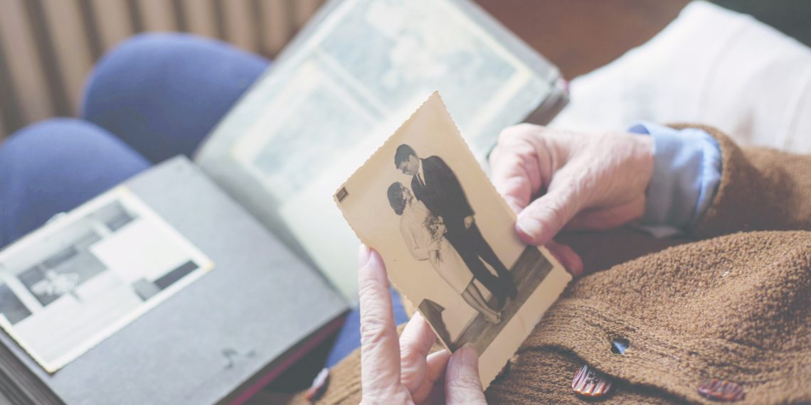 An elderly person holds a vintage photograph of a couple while reminiscing over an open family photo album resting on their lap.