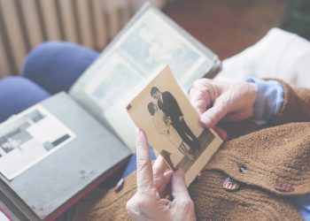An elderly person holds a vintage photograph of a couple while reminiscing over an open family photo album resting on their lap.