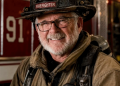 An older firefighter with glasses and a white beard smiles while wearing a black helmet marked “91” and tan firefighting gear in front of a red fire truck.