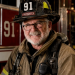 An older firefighter with glasses and a white beard smiles while wearing a black helmet marked “91” and tan firefighting gear in front of a red fire truck.