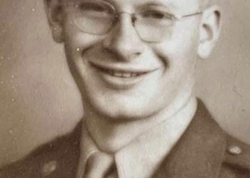 A smiling young World War II U.S. Army soldier wearing glasses, a military uniform, and a service cap poses for a vintage sepia-toned portrait.
