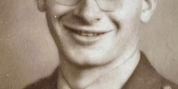 A smiling young World War II U.S. Army soldier wearing glasses, a military uniform, and a service cap poses for a vintage sepia-toned portrait.