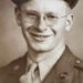 A smiling young World War II U.S. Army soldier wearing glasses, a military uniform, and a service cap poses for a vintage sepia-toned portrait.