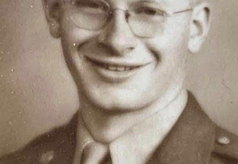 A smiling young World War II U.S. Army soldier wearing glasses, a military uniform, and a service cap poses for a vintage sepia-toned portrait.