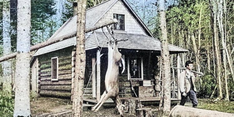 A historic photograph of a log hunting cabin in the woods shows a deer hanging from a tree and a hunter standing outside with a rifle resting on his shoulder.