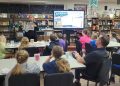Jefferson-Morgan Elementary GATE students listen to a presenter holding a magazine page beside a screen displaying Backroads Magazine in their school library.
