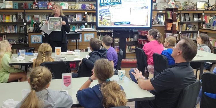 Jefferson-Morgan Elementary GATE students listen to a presenter holding a magazine page beside a screen displaying Backroads Magazine in their school library.