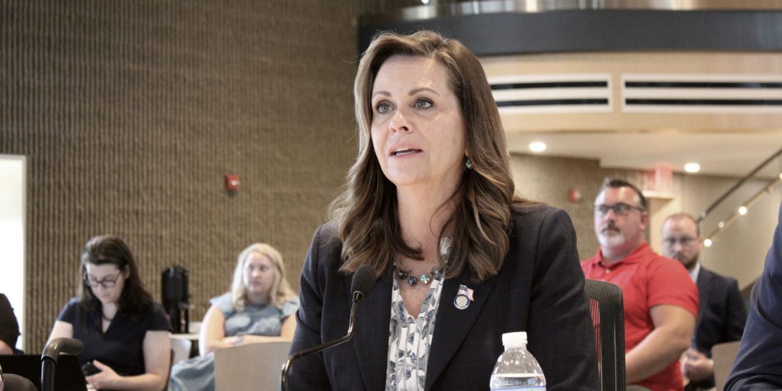Pennsylvania State Senator Camera Bartolotta, wearing a dark blazer and patterned blouse, speaks into a microphone during a public hearing with attendees in the background.
